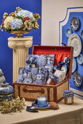A styled gift set arranged in an open vintage-style suitcase on a table, featuring blue-and-white patterned paper goods, blue ribbon bows, a blue candle, and a small jar, set against a blue and cream decorative backdrop with floral arrangements and ceramic plates.